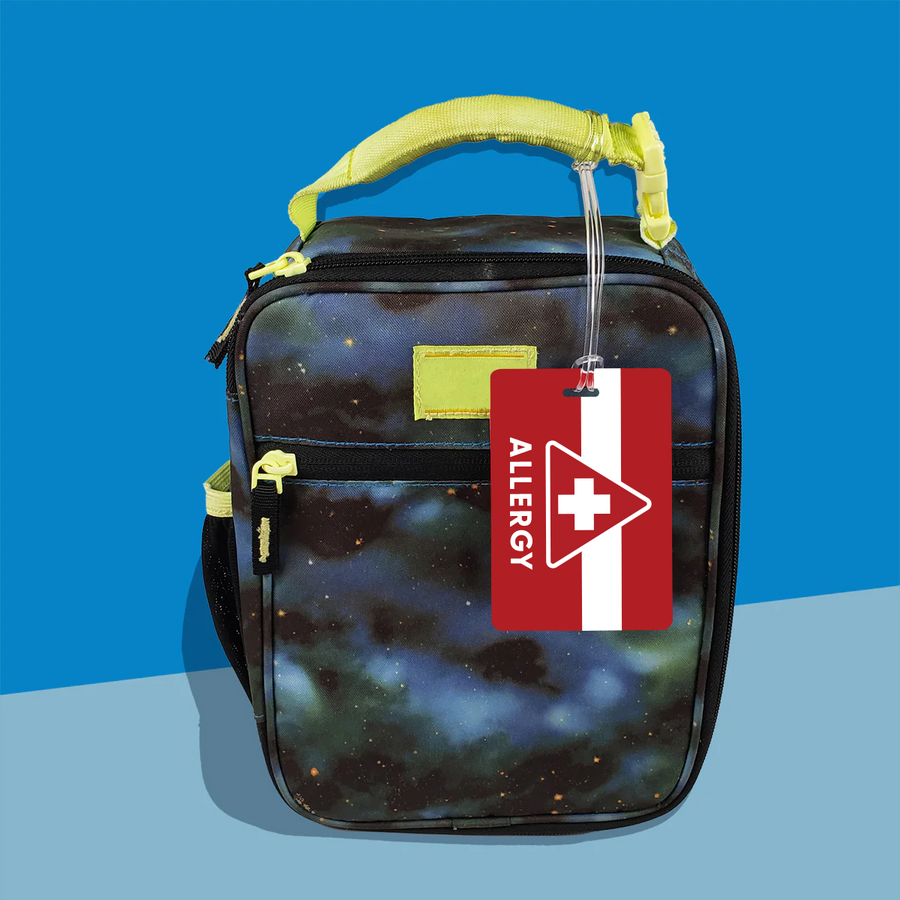 A kids' galaxy-designed lunchbox with a space-themed design, featuring a personalized bag tag with 'ALLERGY' alert and a plastic loop attached to it.