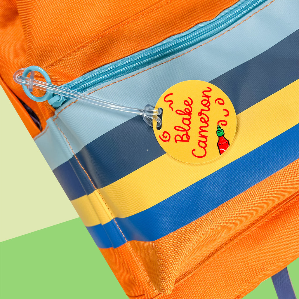 InchBug circular kid's bag tag hanging on an orange bag, text reads "Blake Cameron"