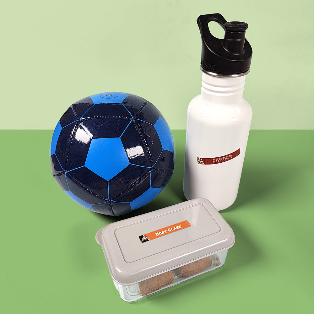 A soccer ball, a water bottle, and a food container with InchBug personalized name labels applied to them, presented on a two-tone green background.