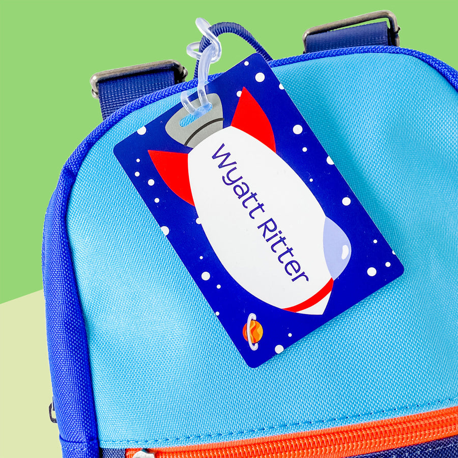 A personalized rectangular InchBug kids bag tag attached to a blue bag, featuring a space shuttle design with the name 'Wyatt' printed on it.