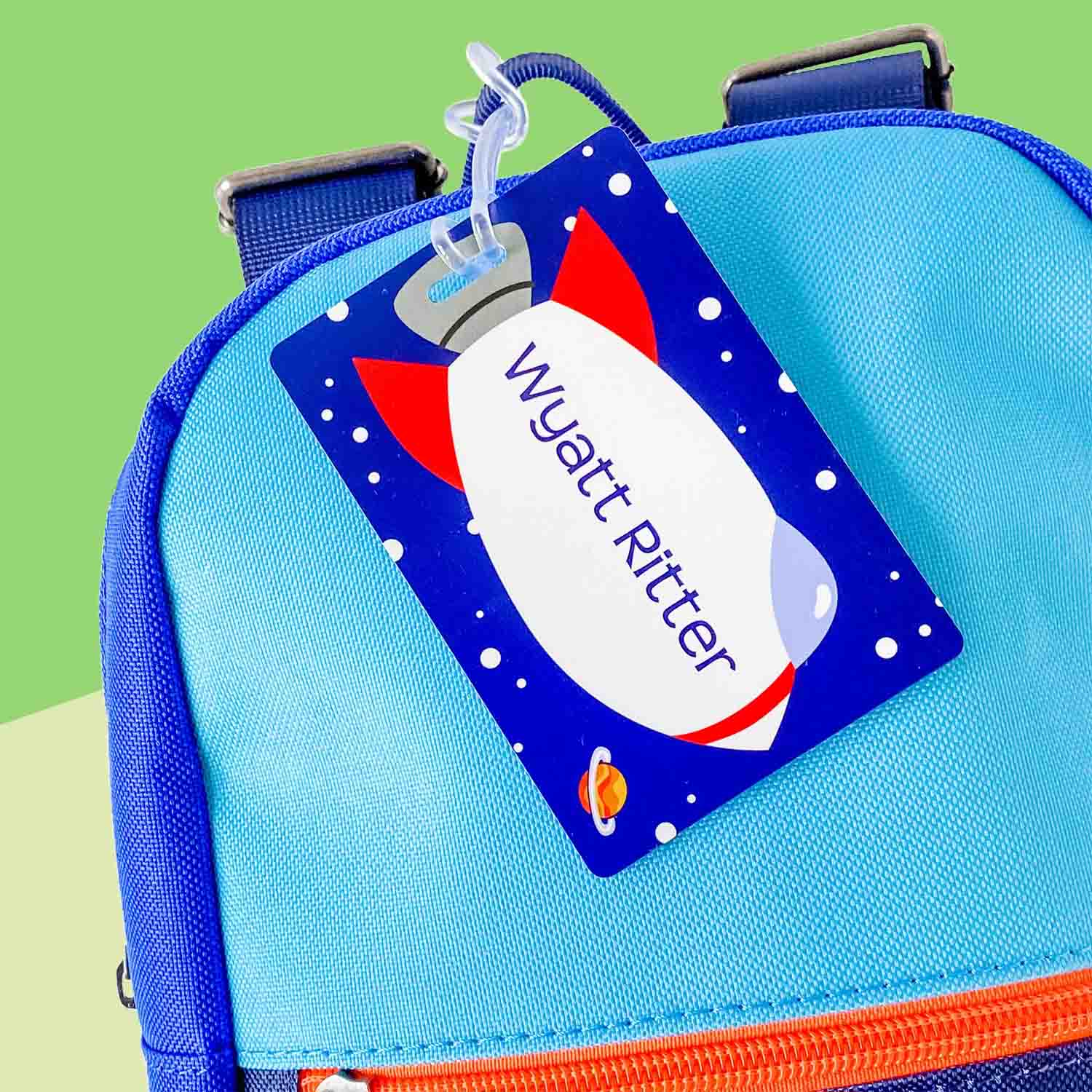 A personalized rectangular InchBug kids bag tag attached to a blue bag, featuring a space shuttle design with the name 'Wyatt' printed on it.