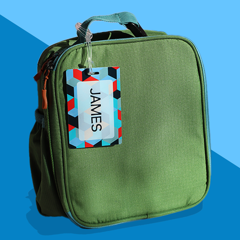 A green kids' bag with a personalized rectangular bag tag attached to it featuring a geometric pattern with the name 'JAMES'.