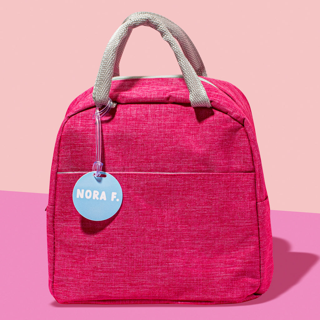 A pink bag with a circular blue bag tag attached to it, with the text 'NORA F.'