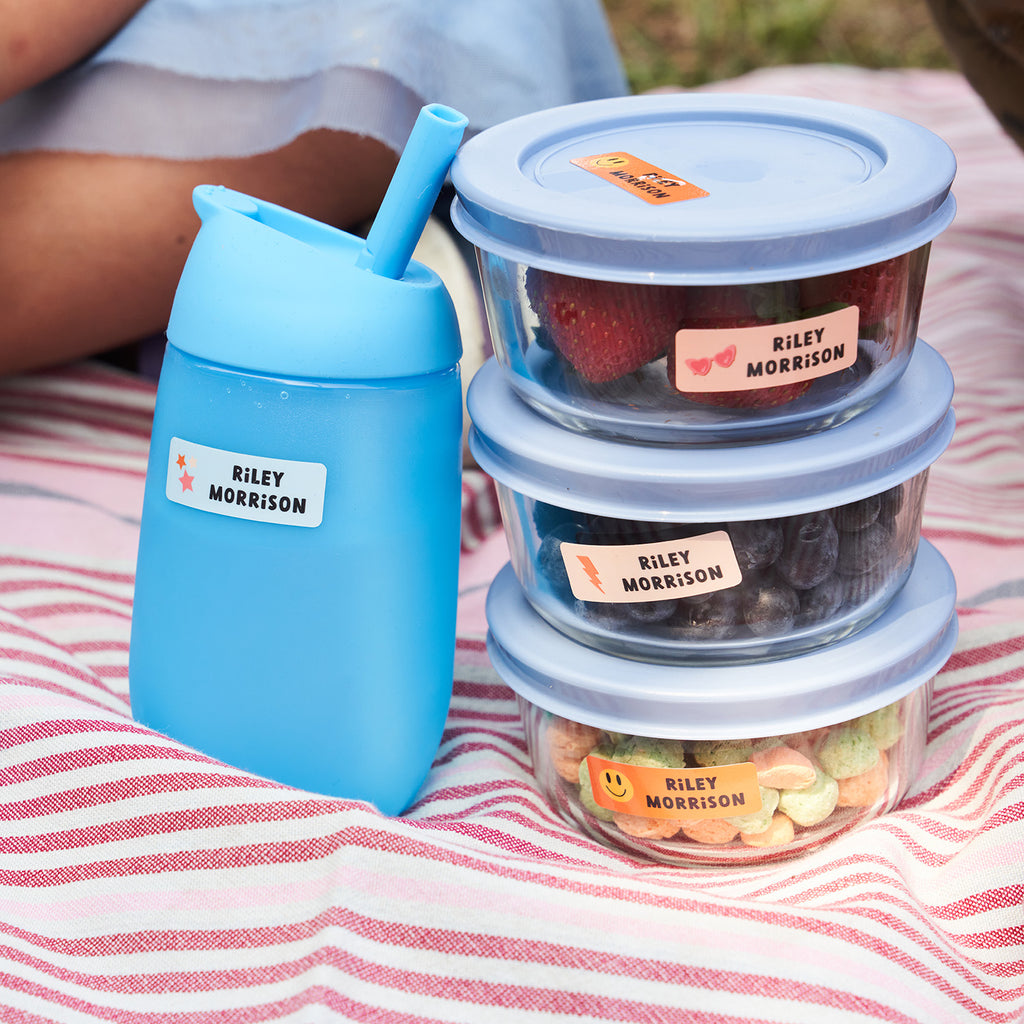 blue water bottle and stacked food containers placed on top of a striped picnic mat, all labeled with rectangular InchBug name stickers for kids