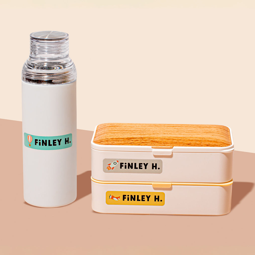 a water bottle and stacked food containers labeled with InchBug personalized stickers for kids, placed against a two-tone beige background
