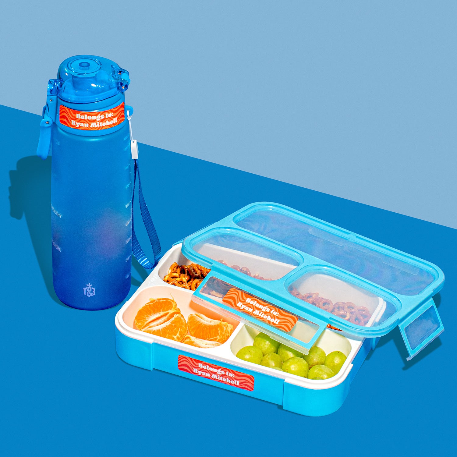 A set of InchBug adhesive name labels on a blue background, with one label applied to a blue water bottle and the others displayed next to a compartmentalized blue lunch box containing fruits and snacks.