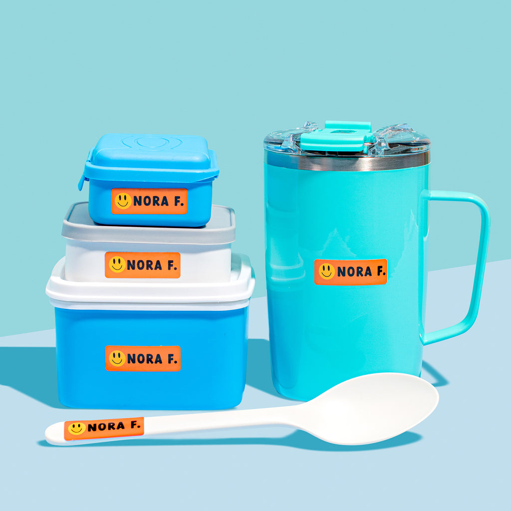 A set of orange InchBug sticker labels for kids on various items including a thermos, lunch box, and a spoon, all personalized with the name NORA F.