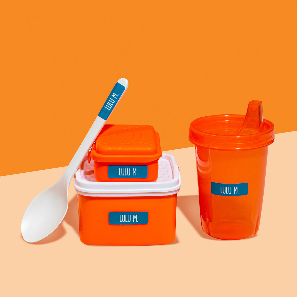 A set of orange food storage containers with white lids, one with a spoon, and all displaying a bold blue personalized InchBug sticker labels for kids