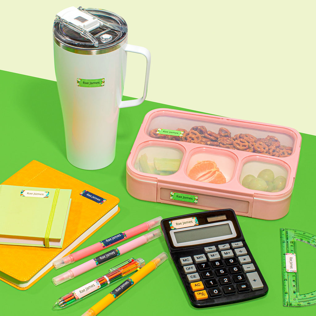 A collection of school supplies including a lunch box, a thermos, notebooks, a calculator, pens, and a ruler, all laid out on a table, all labeled with various colors of InchBug sticky name labels