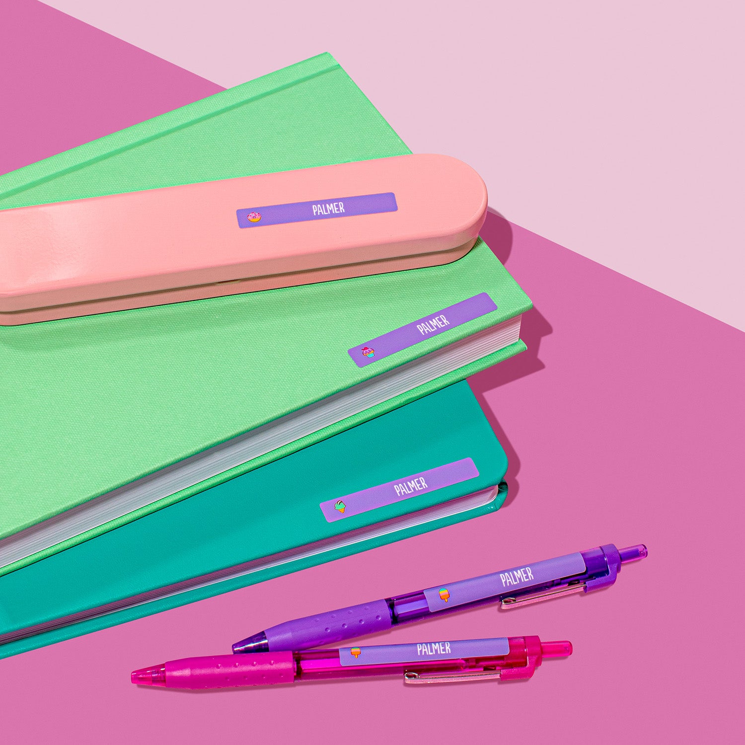 Purple InchBug name labels placed on a surface with some labels on top of colored notebooks and pink pencil case, and two pens lying beside them.