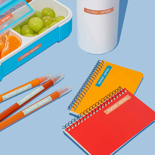 InchBug mini sticker name labels placed on several items, including food container, water bottle, pens, and notebooks