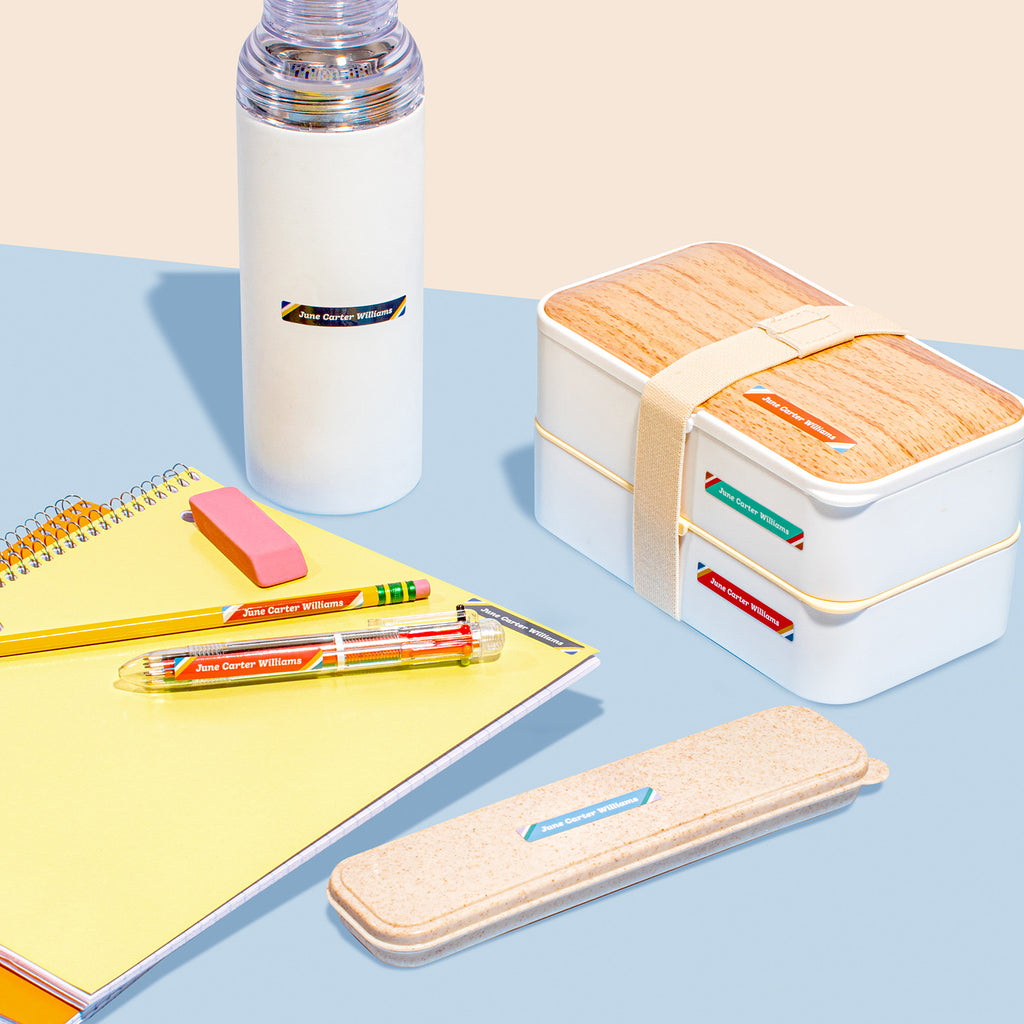 A collection of items including a water bottle, a lunch box with wooden accents, a notebook, pens, and a ruler, all laid out on a two-tone background, labeled with InchBug Mini Rectangle Name Labels - Teen Line.