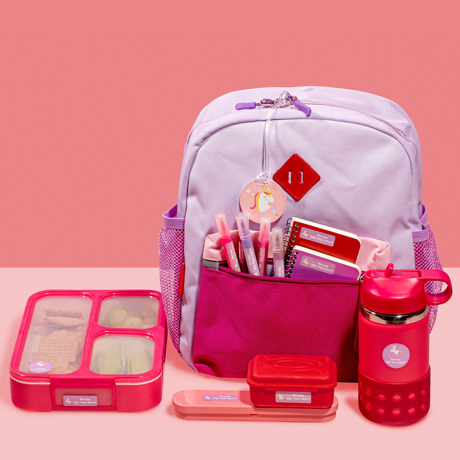a purple backpack, red water bottle, red food container, and other items labeled with the InchBug labels and bag tag on a two-toned pink and beige background