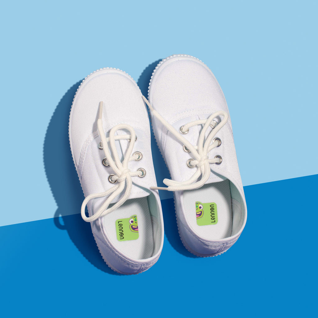 A pair of white sneakers on a two-tone blue background, featuring green InchBug shoe labels attached to the soles.