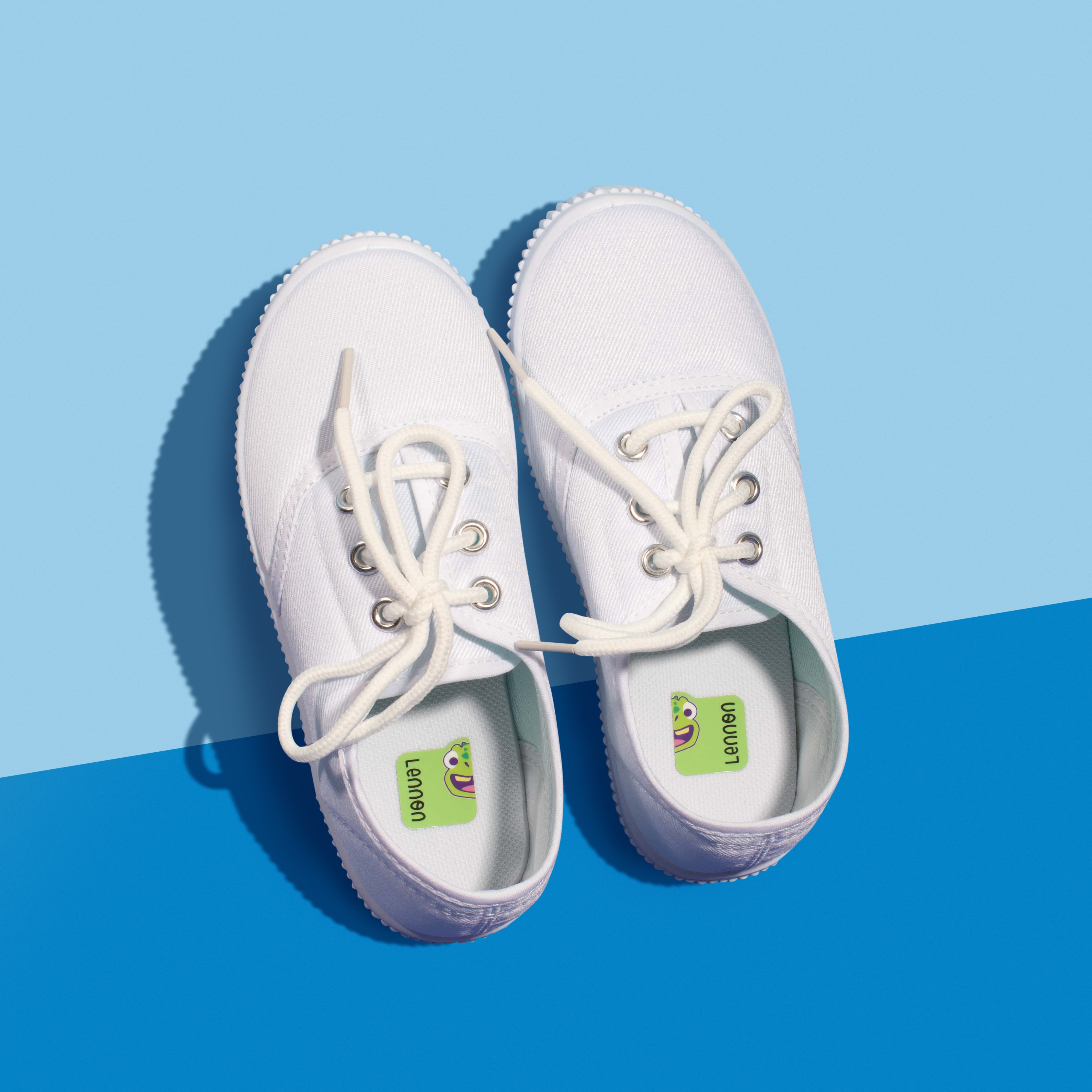 A pair of white sneakers on a two-tone blue background, featuring green InchBug shoe labels attached to the soles.