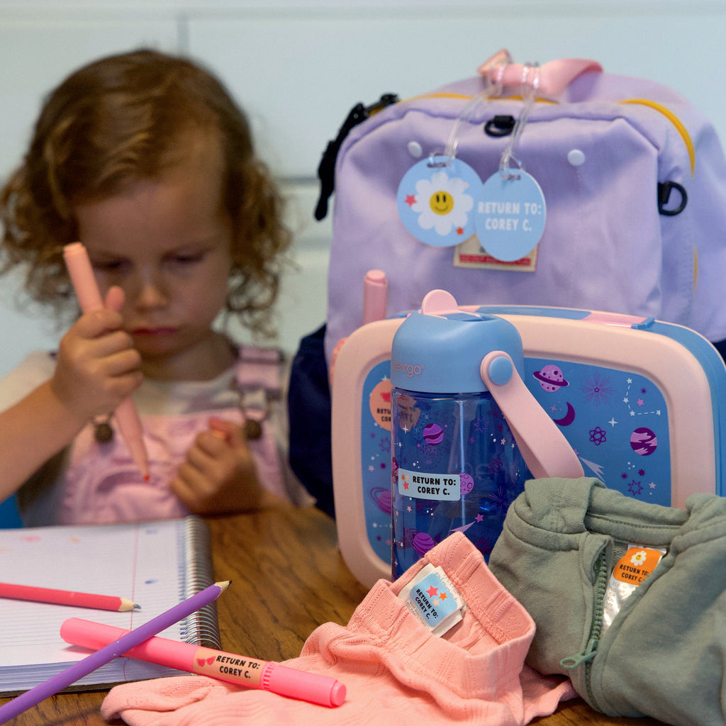 clothes, backpack, and other essentials labeled with InchBug labels and bag tag, laid in front of a kid holding a pink pen