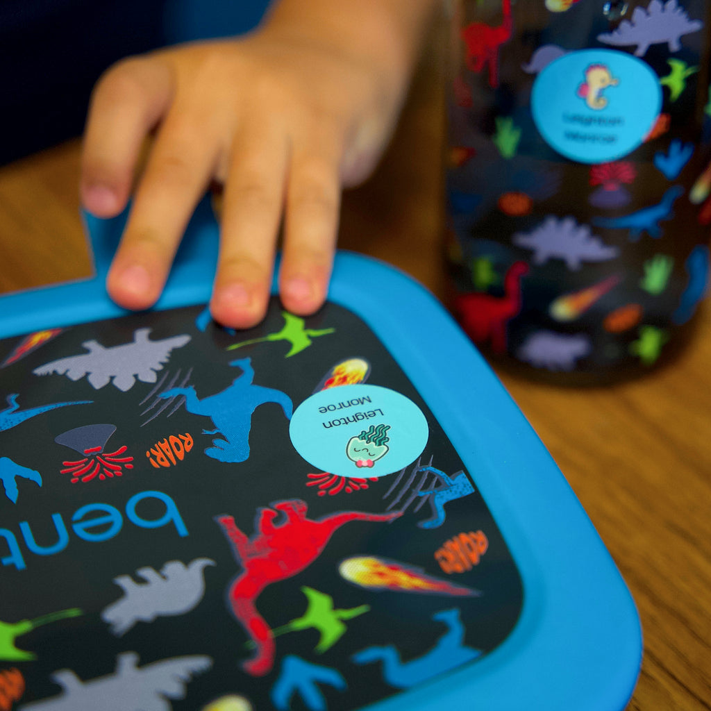 kid placing their hand on a food container labeled with a blue InchBug sticker label for kids; out of focus is a water bottle with dinosaur prints labeled with a blue InchBug label