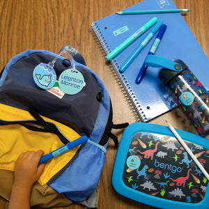 a child's hand holding a blue pen labeled with an InchBug name sticker, alongside other essentials like a backpack, lunch box container, water bottle, and a notebook, all also labeled with InchBug labels