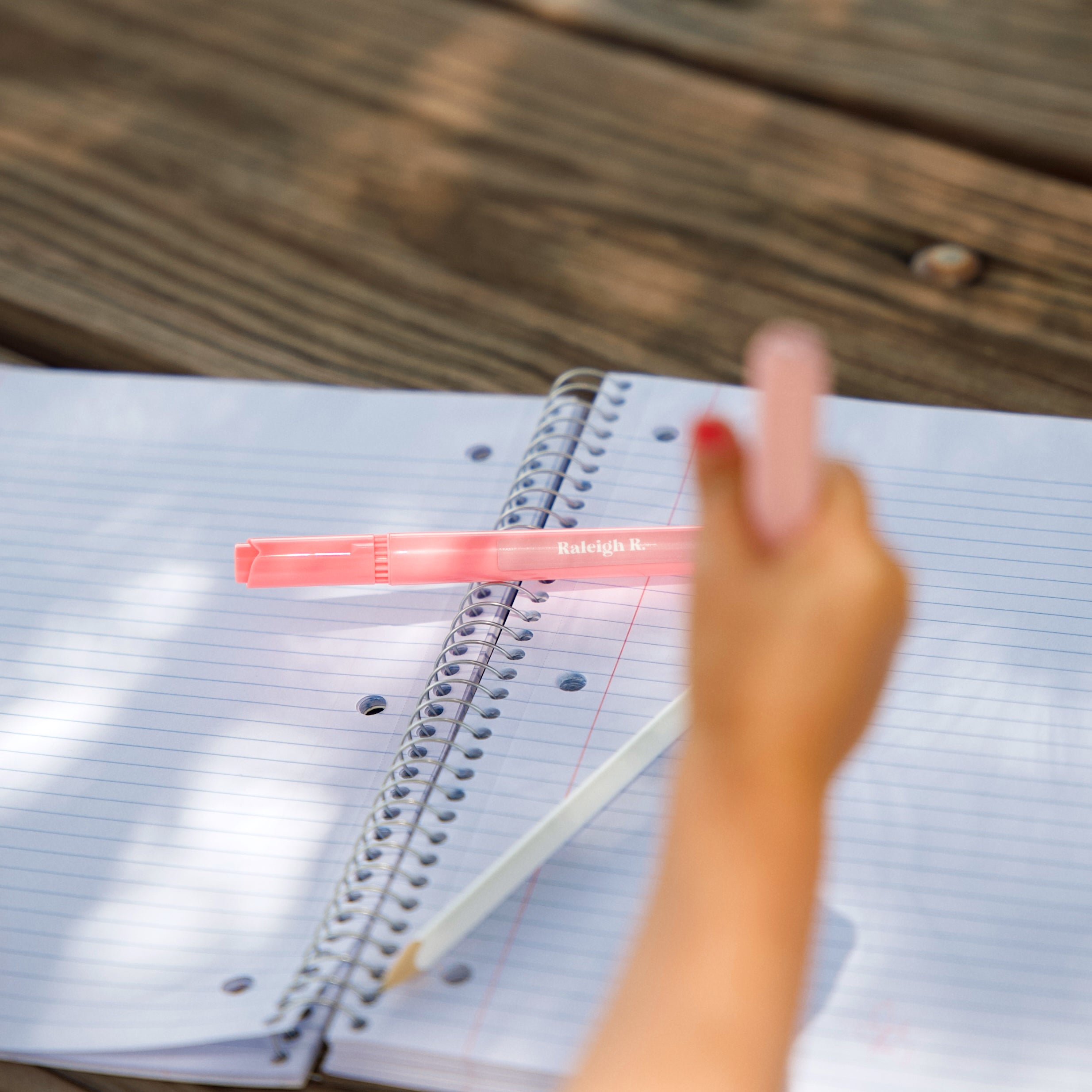 an open notebook with a pen and pencil on top of it; out of focus is a child's hand holding a pink pen