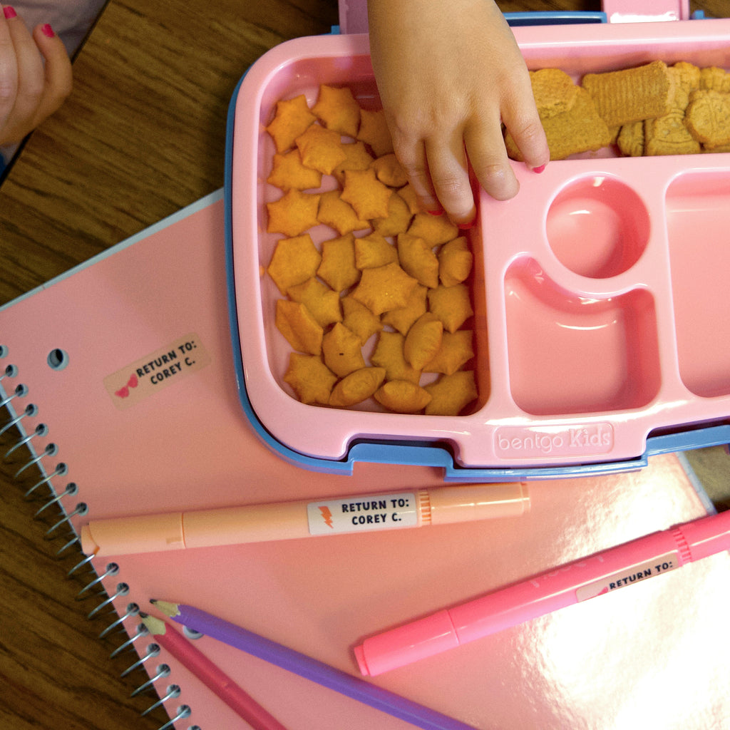child's hand getting a biscuit on a pink container placed on top of a pink notebook also with pens on top of it, all labeled with sticky name tags that read "RETURN TO: COREY C."