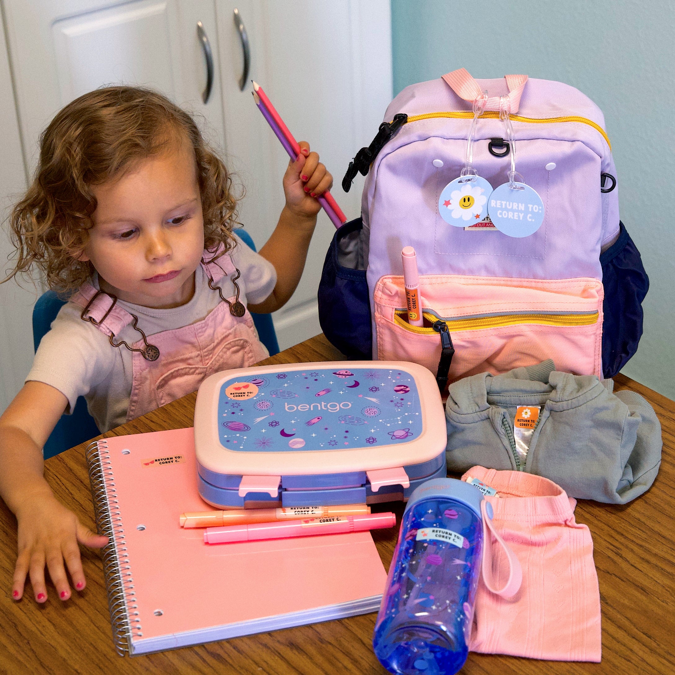A child holding pencils, surrounded by items labeled with InchBug's Back To School Combo Pack, which are good personalized name labels for school.