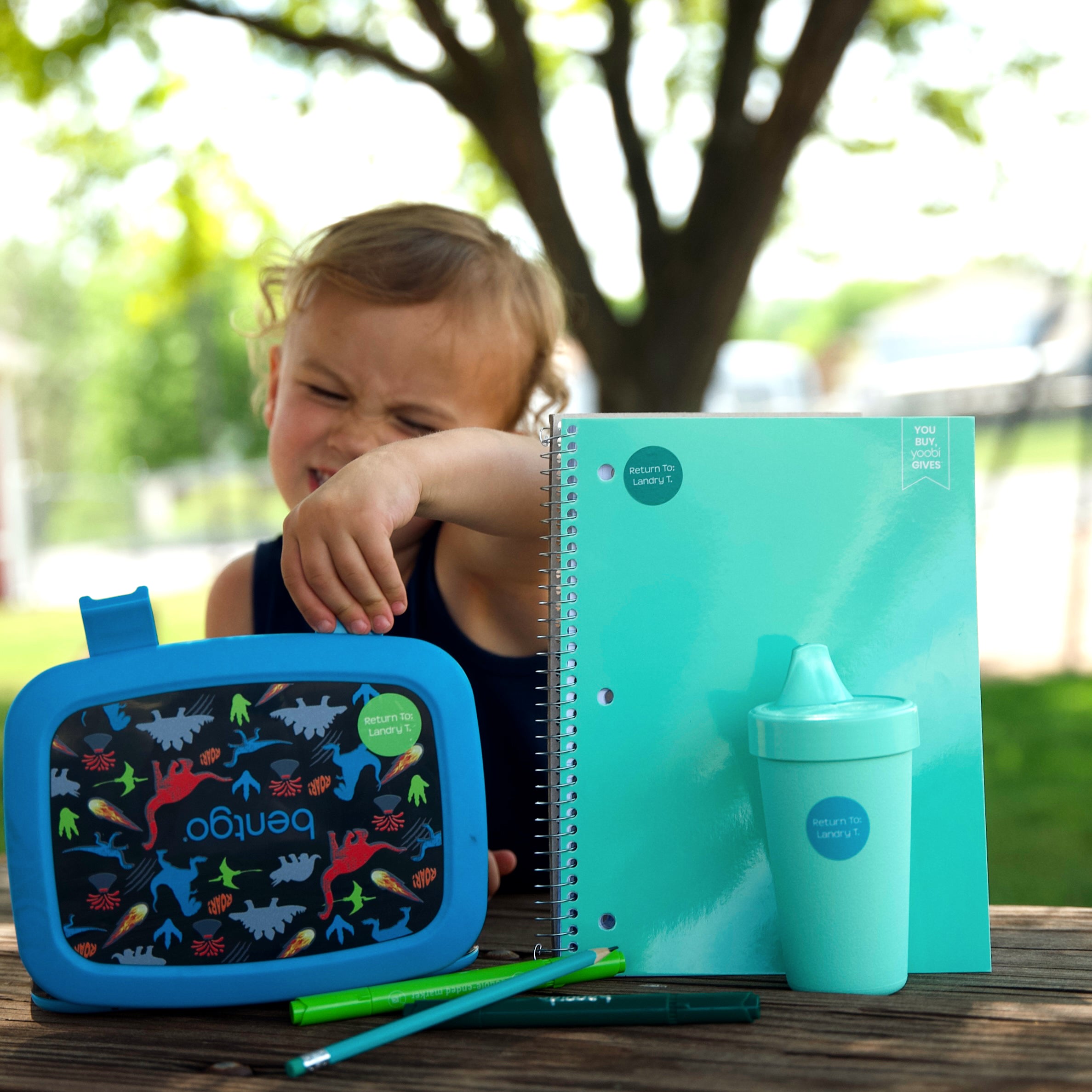 a kid posing behind school essentials like a green notebook, a green water container, a blue food container, and pens and pencils, all labeled with InchBug labels